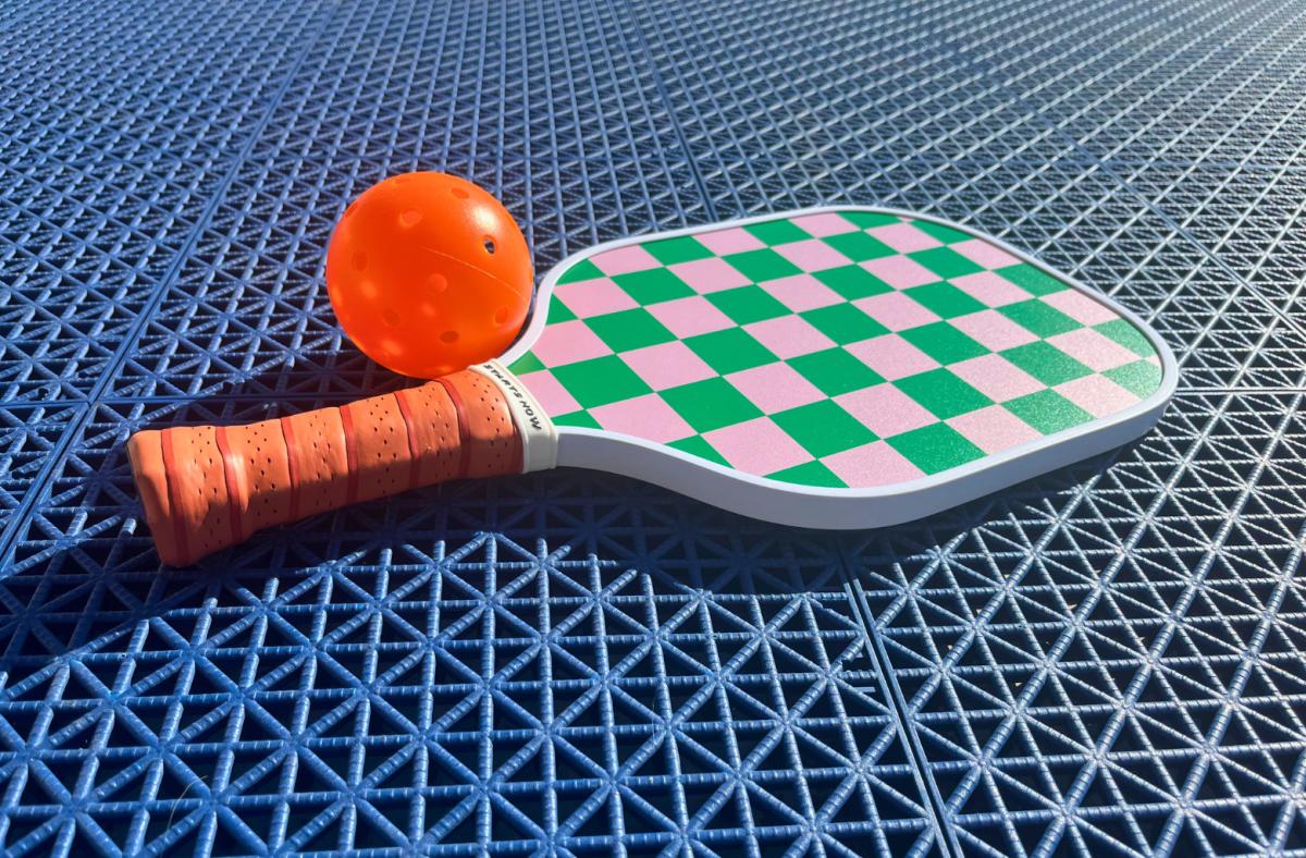 On Holiday pink and green checkered pickleball paddle with an orange handle on a blue textured surface next to an orange ball.